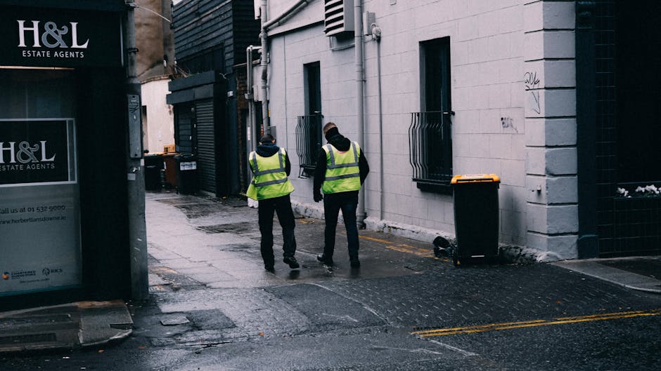 Two maintenance workers wearing high-visibility yellow vests and dark clothing stand on a wet, paved alleyway between buildings in an urban area. One worker is holding a spray bottle, while the other