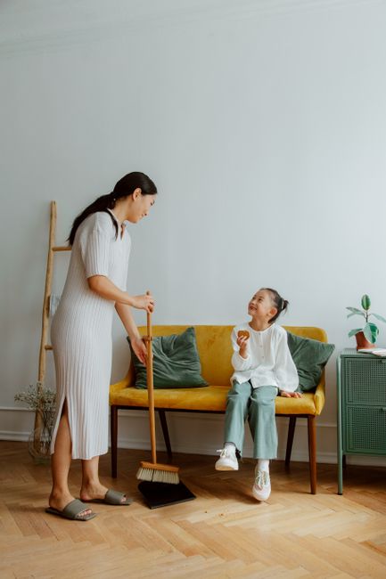 A woman with long dark hair, dressed in a white knitted dress, is standing indoors holding a broom in her right hand, engaging in surface cleaning. She is facing a young girl sitting on a yellow sofa with green cushions, who is smiling and holding a snack. The room features a light-colored wall, a wooden floor, and a small green side table with a potted plant. In the background, there is a wooden ladder-style shelf leaning against the wall and a lamp on the side table, indicating a cozy living space. The scene emphasizes domestic cleaning and tidying activities, highlighting the importance of proper surface maintenance and hygiene, aligning with the theme of professional quality cleaning services offered by Ilford Carpet Cleaners.