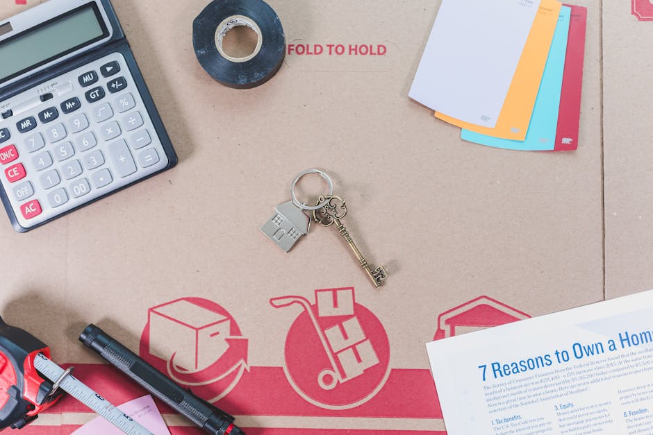 A flat lay image featuring a beige surface with various items related to home and office organization. Visible are a black calculator with white and pink buttons, a roll of black tape, a group of colorful index cards, a set of keys including a silver house key, a small silver house-shaped keychain, and a silver keyring. Additionally, there is a black marker, a pink measuring tape, and a document titled '7 Reasons to Own a Home' with blue text. In the background, there is a cardboard packing sheet with red 'FOLD TO HOLD' text and symbols indicating packaging instructions. The setting appears to be a workspace or organization area, emphasizing cleanliness and preparation, with natural lighting highlighting the neat arrangement of items. For surface cleaning and sanitisation of household or office spaces, Ilford Carpet Cleaners offers professional domestic cleaning services.