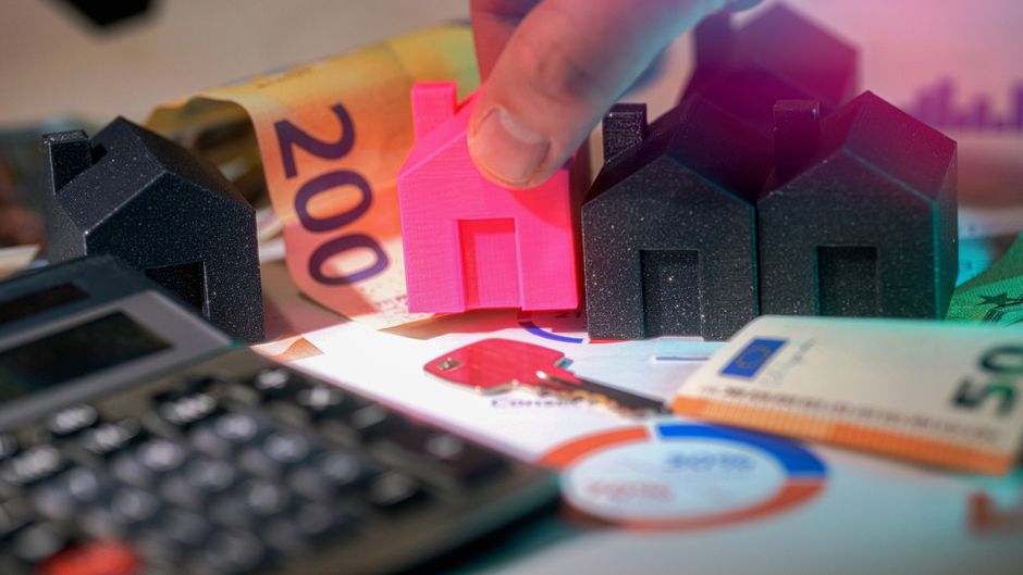 Close-up image showing a person's hand holding a small pink house model among black and pink house figurines on a surface that includes a calculator, financial papers, and currency notes, illustrating financial or property management. The background features a rolled-up banknote and a green paper currency note, suggesting financial calculations related to property or real estate. The scene is well-lit, emphasizing the details of the objects, with a focus on the miniature house models and monetary elements, aligning with themes of property costs or financial planning connected to end of tenancy cleaning services. The image conveys a professional and organized atmosphere suitable for content about domestic or commercial cleaning costs, as discussed in the Ilford End of Tenancy Cleaning Real Cost Guide, provided by Ilford Carpet Cleaners.