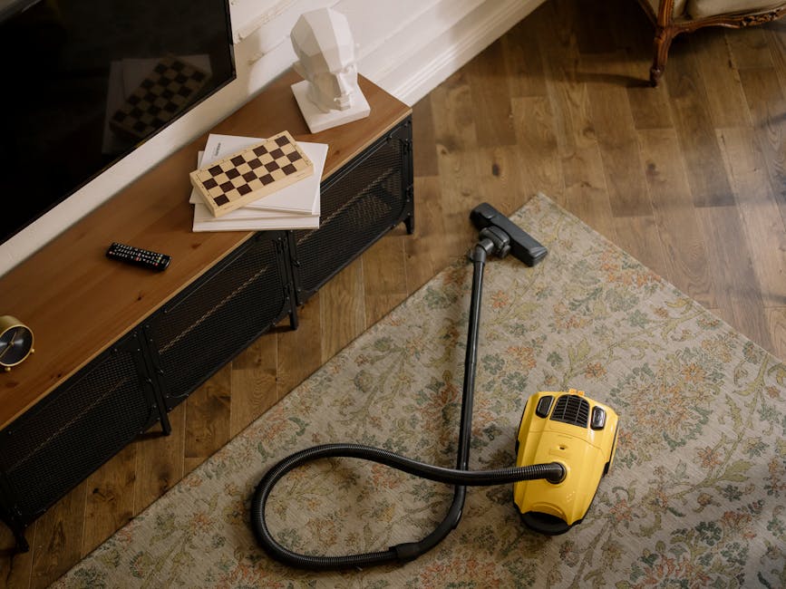 A yellow canister vacuum cleaner with a black hose and silver wand attachment is placed on a patterned area rug on a wooden floor in a living room. The rug features a light, intricate floral design. To the left, there is a wooden media console with black metal legs, topped with a white bust sculpture, a remote control, and a small stack of papers or books. The room is illuminated by natural light, and a brown upholstered chair is partially visible in the background. The scene reflects a clean, well-maintained domestic space, as part of a professional cleaning service by Ilford Carpet Cleaners featured on the page for same-day carpet cleaning and instant quotes.