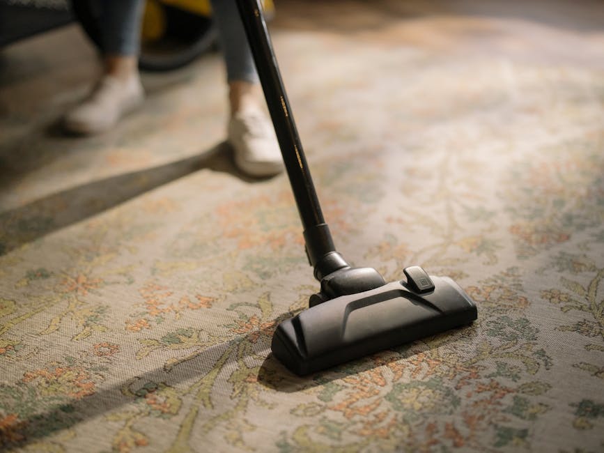 A yellow canister vacuum cleaner with a black hose and silver wand attachment is placed on a patterned area rug on a wooden floor in a living room. The rug features a light, intricate floral design. To the left, there is a wooden media console with black metal legs, topped with a white bust sculpture, a remote control, and a small stack of papers or books. The room is illuminated by natural light, and a brown upholstered chair is partially visible in the background. The scene reflects a clean, well-maintained domestic space, as part of a professional cleaning service by Ilford Carpet Cleaners featured on the page for same-day carpet cleaning and instant quotes.
