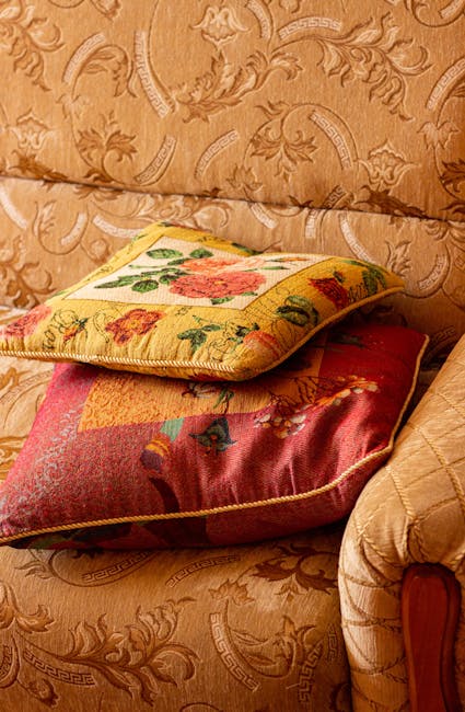 Close-up of two decorative cushions resting on a patterned fabric sofa in a living room. The top cushion features a floral embroidery design with pink and green accents on a yellow background, while the bottom cushion has a red and orange quilted pattern. The sofa's surface is covered with a beige or light brown fabric that has a subtle, ornate embroidered or printed pattern. The lighting is soft and natural, highlighting the textures and colors of the cushions and upholstery, with no visible dust or stains. This image visually illustrates domestic surface care and upholstery cleaning, aligning with the page title 'Seven Kings Upholstery Cleaning Access Problems Solved' on ilfordcarpetcleaners.com, as part of their comprehensive cleaning and sanitisation services.