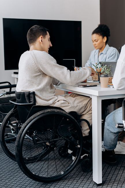 A person in a wheelchair is seated at a white table in a modern office or meeting room, engaging in conversation with a woman sitting across from them. The table holds a potted plant and some papers or notebooks. Behind them, a large flat-screen monitor is mounted on a white wall, illuminated by soft lighting. The room features a grey carpeted floor, and the overall environment appears clean, organized, and well-maintained. The setting suggests a professional space focused on accessibility, communication, and community engagement, aligning with the themes of surface cleaning and hygiene as part of comprehensive facilities management promoted by Ilford Carpet Cleaners.