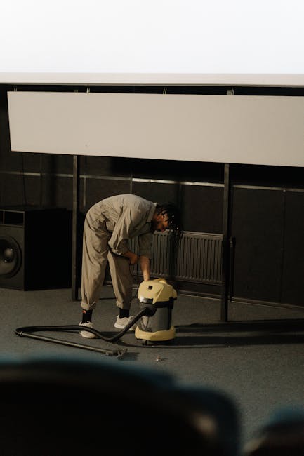 A person in light-colored overalls is engaged in deep cleaning or surface cleaning activities within a room, using a beige vacuum or carpet cleaning machine. The room features a dark wall with a white horizontal panel or radiator and a black speaker in the background. The floor appears to be carpeted, and the lighting is bright, highlighting the cleanliness process. This image demonstrates professional domestic or commercial cleaning, associated with Ilford Carpet Cleaners, correlating with the Valentines Park Carpet Cleaning Job Before and After page, emphasizing thorough sanitisation and maintenance of carpeted surfaces.